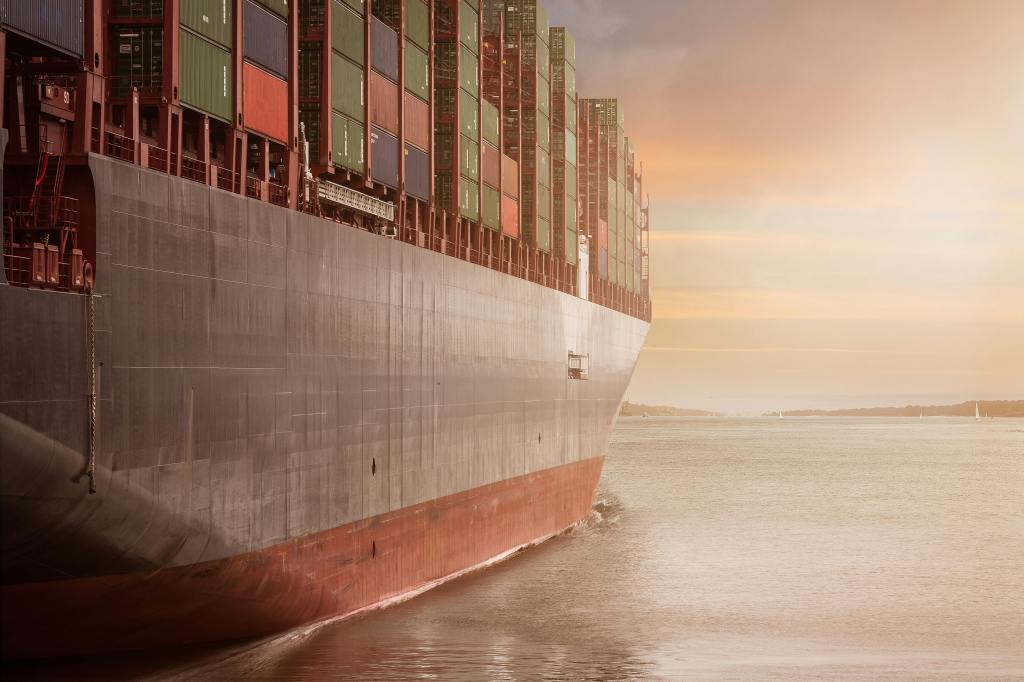 A large cargo ship with colorful containers on its deck sailing on a calm waterway during sunset.