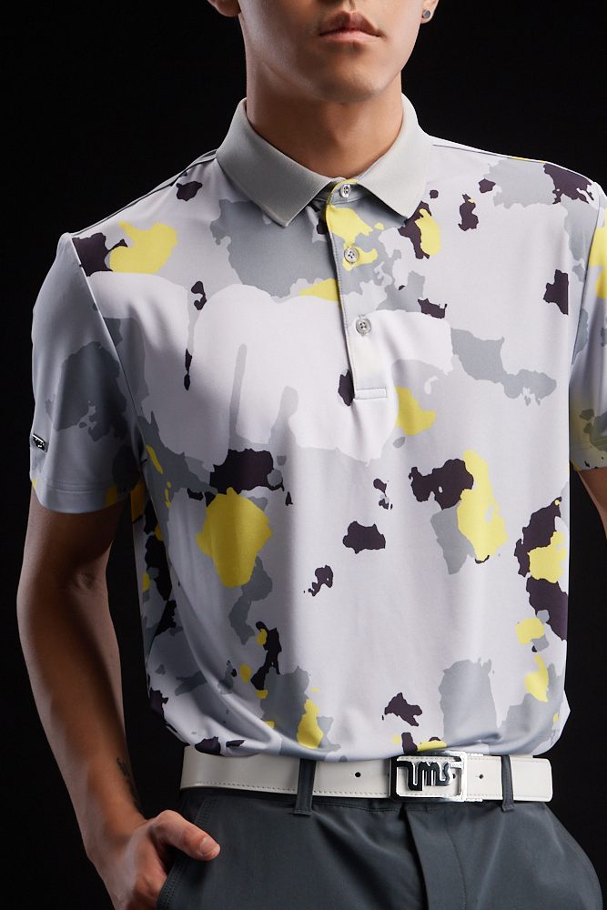 A close-up of a model wearing a patterned polo shirt in shades of gray, yellow, and black, paired with a white belt.