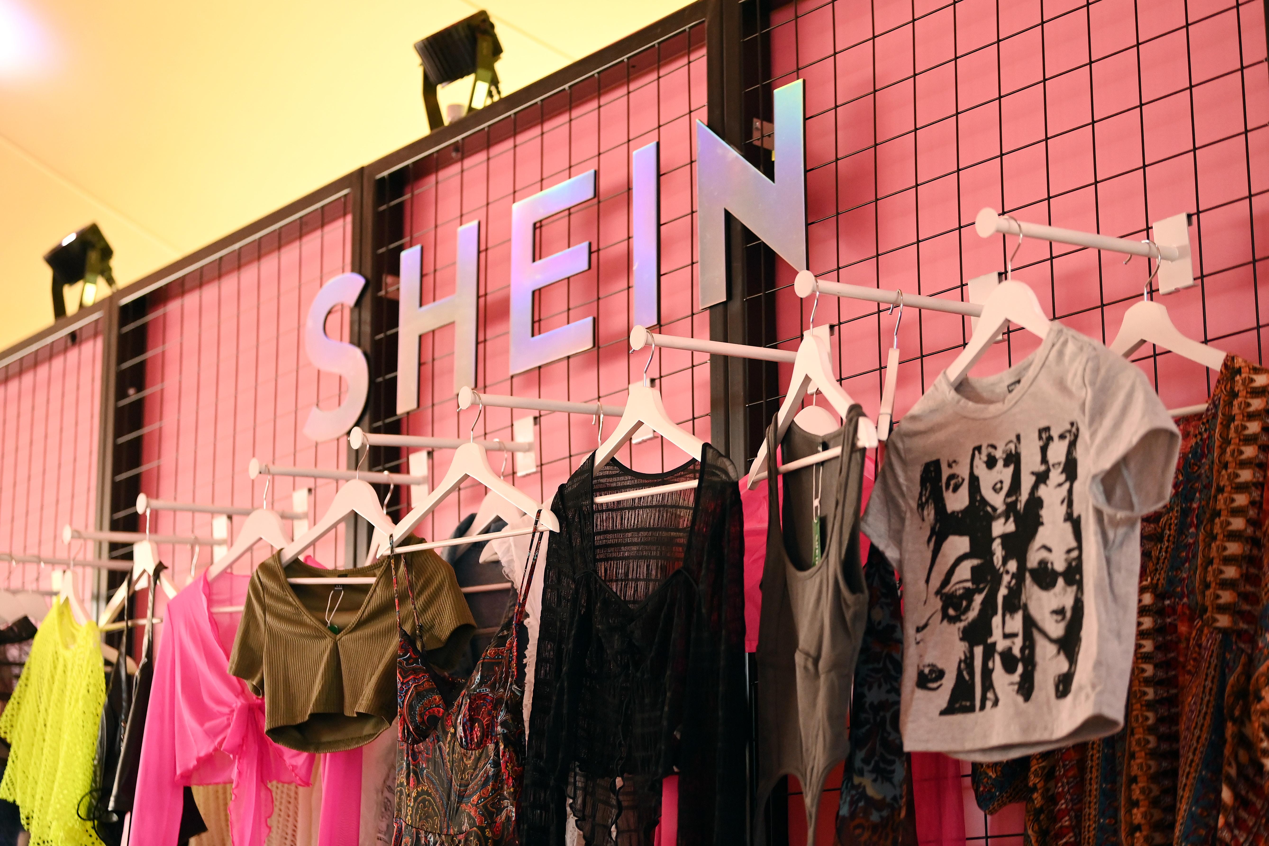 A display of clothing items hanging on white hangers against a pink backdrop, featuring the brand name 'SHEIN' prominently displayed above.