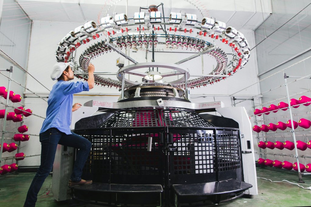 A textile worker operates a high-tech knitting machine in a factory setting, with vibrant pink spools of yarn in the background.