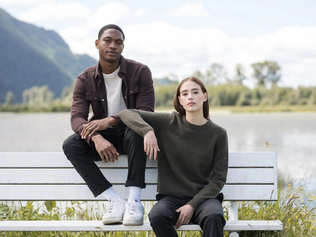 A man and a woman sit on a white bench by a serene lake, surrounded by lush greenery and mountains. The man is wearing a brown button-up shirt over a light-colored top, while the woman is dressed in a dark green sweater and black pants. They both have a relaxed demeanor, with the woman resting her arm on the man's leg.