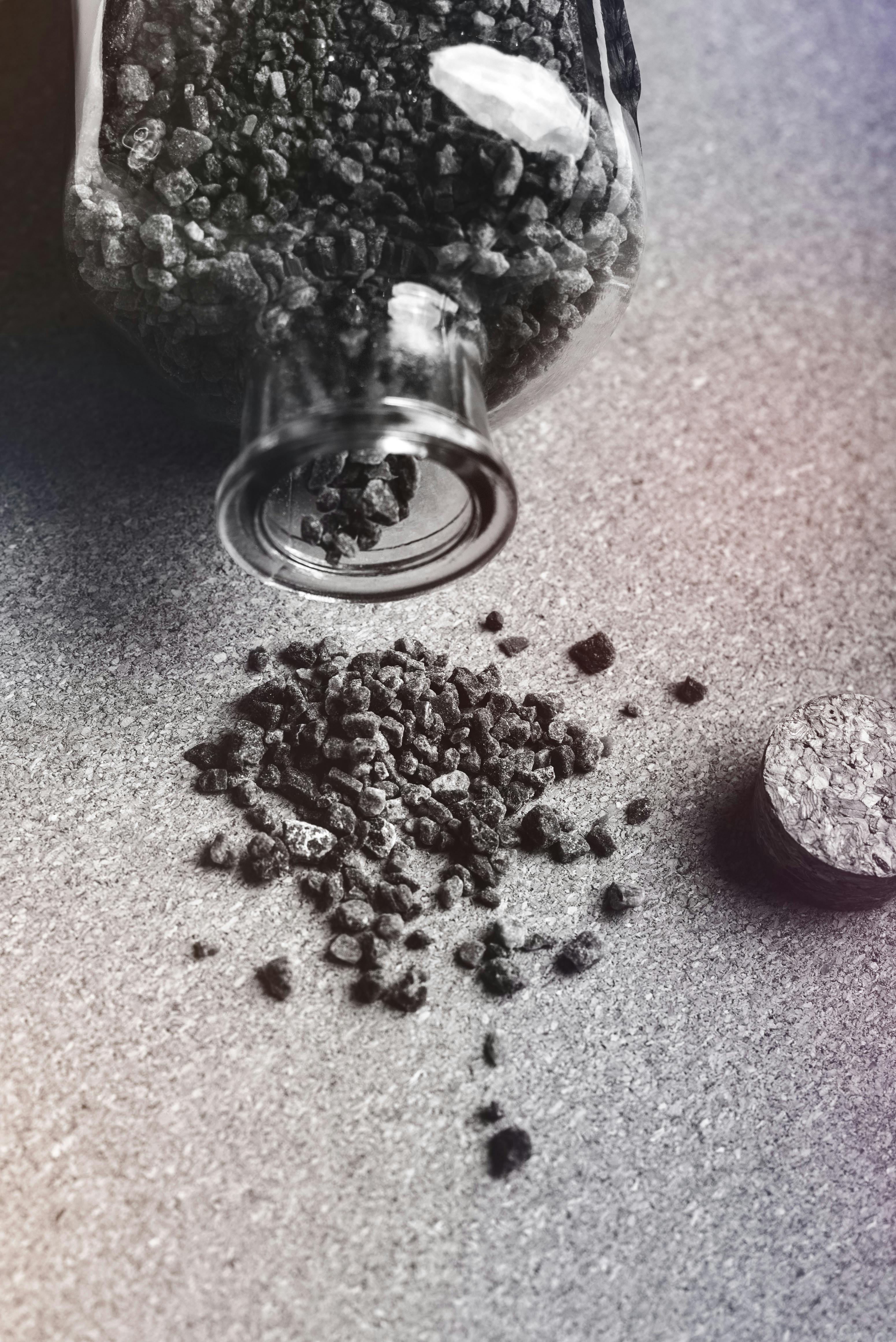 Close-up of a glass jar spilling dark granules onto a textured surface, with a cork lid nearby.