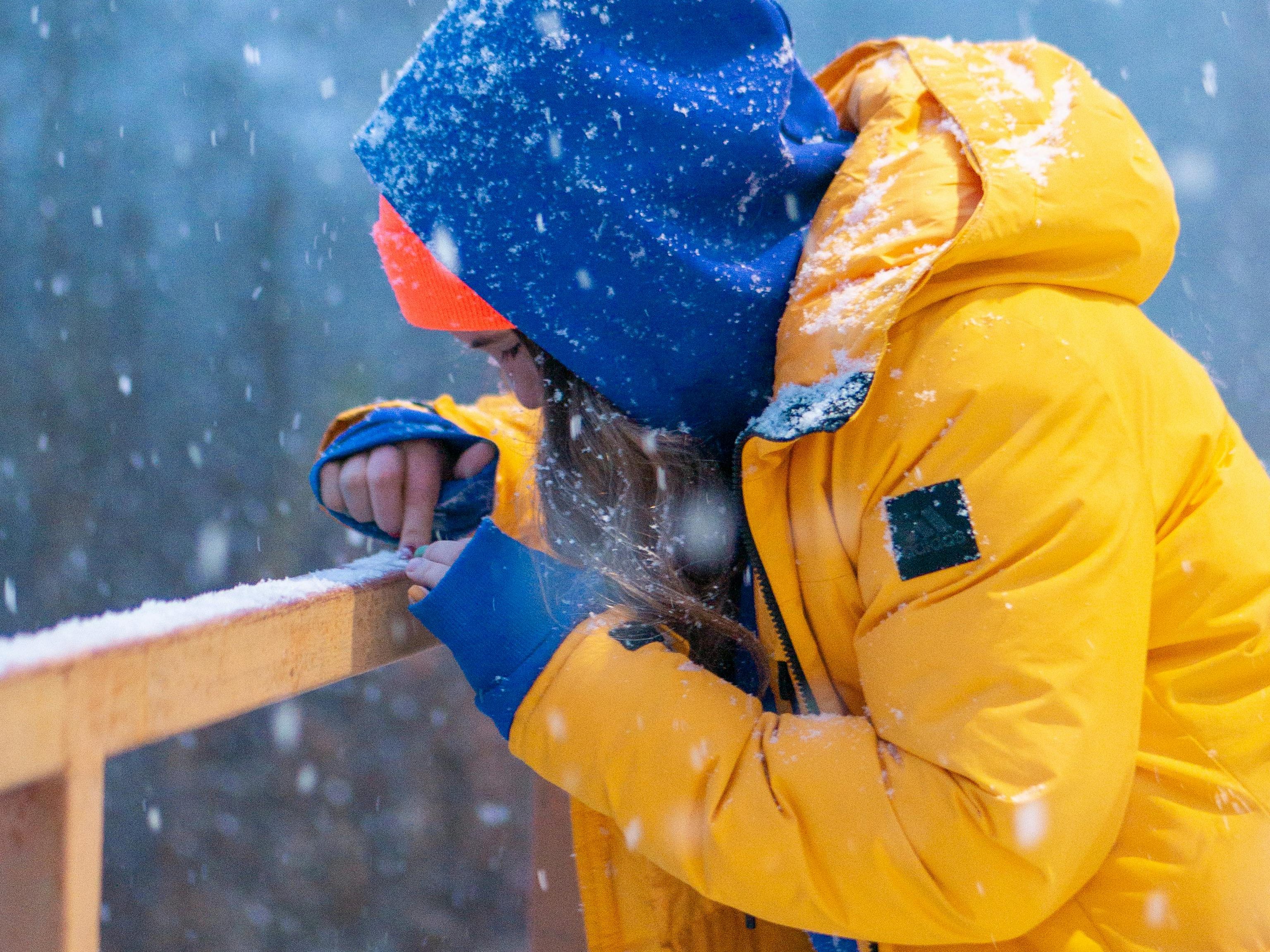 A person wearing a yellow and blue winter jacket, adjusting something on a wooden railing in a snowy environment, demonstrating outdoor durability.