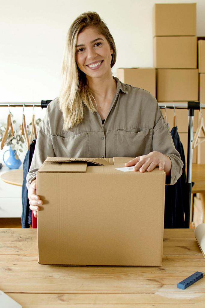 A smiling woman holding a cardboard box, standing at a table in a workspace with clothing items and packing materials in the background.