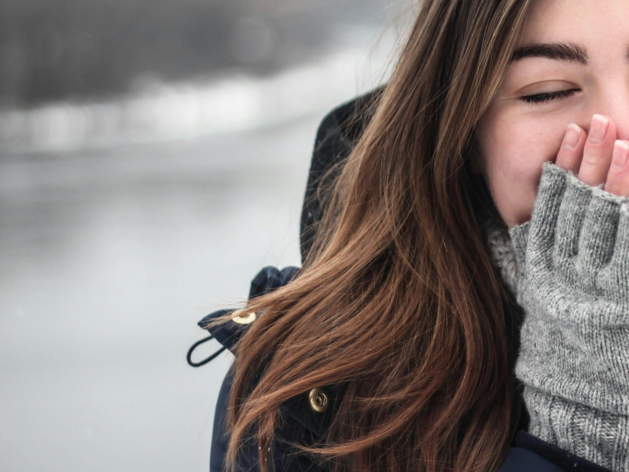 A close-up of a smiling woman wrapped in a warm scarf, with her hands covering her face against a cold outdoor backdrop.