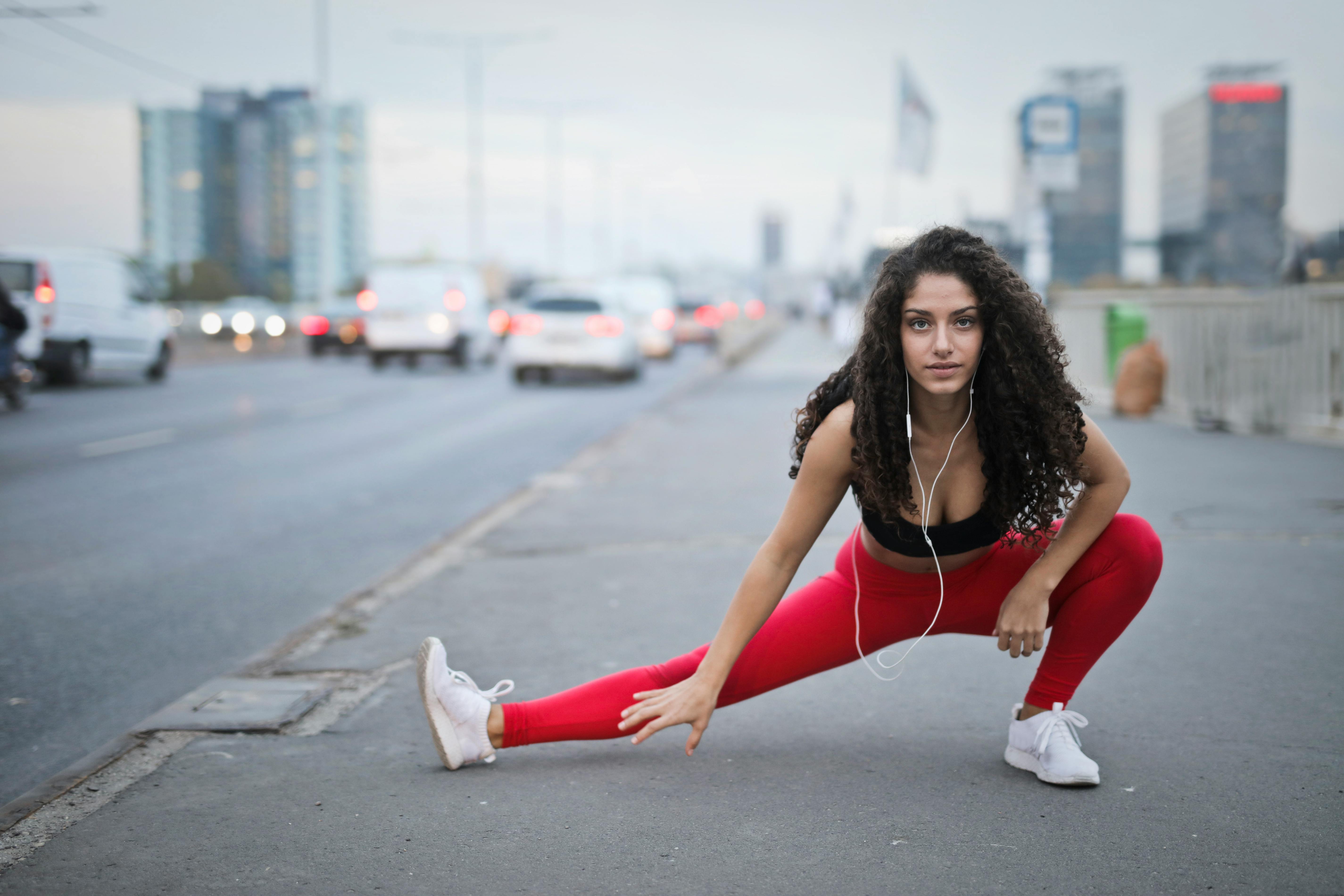 A woman stretching her leg on a city street while wearing athletic wear, with vehicles visible in the background.