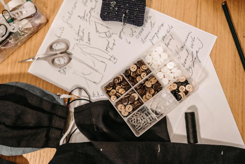 A workspace with fabric samples, scissors, and a container of various buttons, accompanied by a sheet of sketch paper with clothing designs.