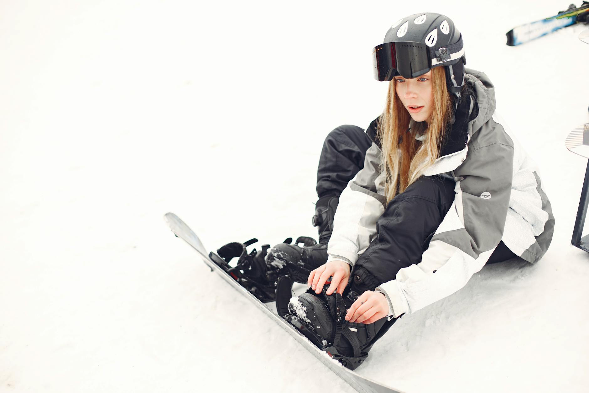 A person sitting on snow, fastening their snowboard boots while dressed in a gray and black ski outfit and helmet.