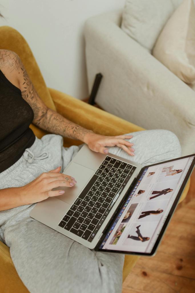 A person is sitting on a yellow chair, wearing a black tank top and gray pants, using a laptop to browse images of clothing.