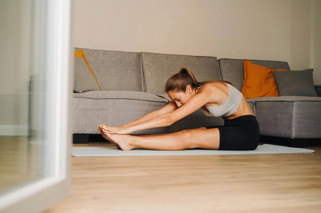 A woman wearing athletic clothing performs a forward stretch on a yoga mat in a living room, with comfortable furniture in the background.