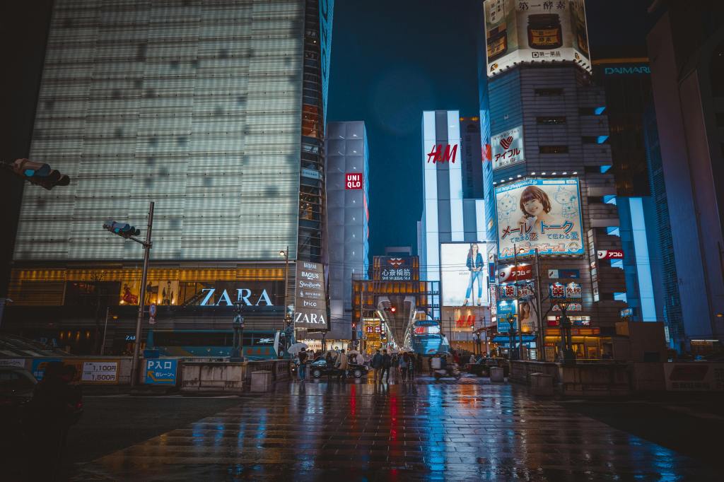 A bustling city street at night, featuring large illuminated storefronts for brands like Zara and H&M, surrounded by vibrant advertisements and reflections on wet pavement.